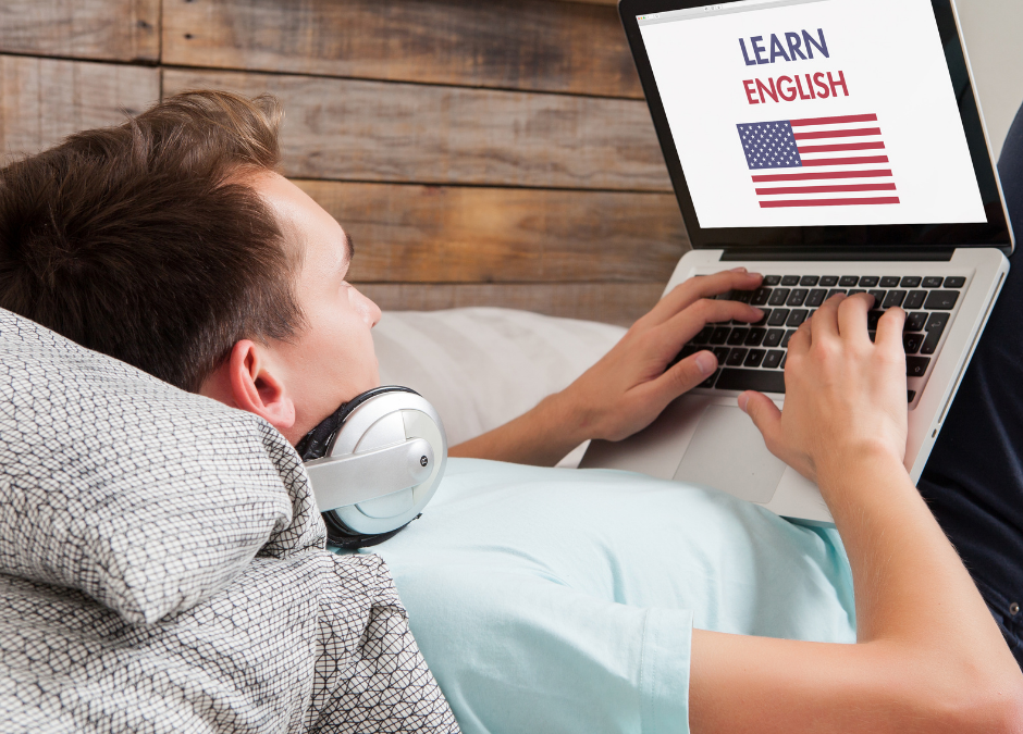 Zdjęcie młodego mężczyzny, który leży na kanapie ze słuchawkami na uszach. Trzyma laptopa na którym widoczna jest grafika – amerykańska flaga, a wyżej napis learn english.